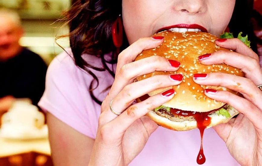 A person with red nail polish takes a big bite of a sesame seed hamburger and tries to curb their food cravings. Ketchup drips from the burger. The person is wearing a pink shirt and red earrings. Another person can be seen blurred in the background. MyFitnessPal blog