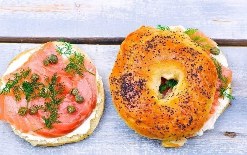 A bagel sandwich on a wooden surface. One half is open, topped with smoked salmon, fresh dill, capers and cream cheese. The other half has poppy seeds on top and partially covers the filling. MyFitnessPal blog