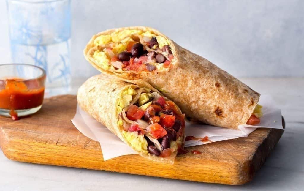 Two burritos filled with scrambled eggs, black beans, tomatoes and cheese are placed on a wooden board covered with baking paper. For those who enjoy varied breakfast options, these burritos are a delicious addition to your 5-egg repertoire. A small glass container of red sauce and a decorative drinking glass set the scene in the background. MyFitnessPal blog