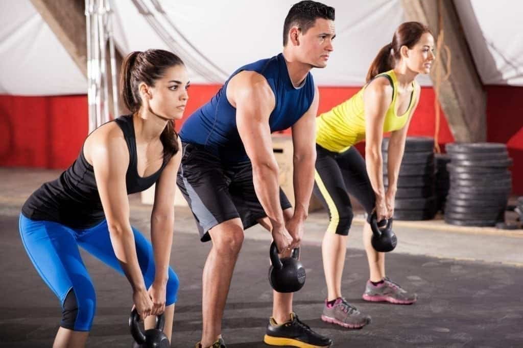 Three people participate in a kettlebell workout at a gym, seamlessly mixing weighted training into their routines. They are lined up side by side and perform a lifting exercise. Two women are wearing athletic tops and leggings, while the man in the middle is wearing a sleeveless top and shorts. MyFitnessPal blog