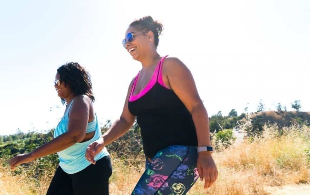 Two women wearing activewear walking outdoors on a sunny day and embracing their walking decisions. One woman wears a blue tank top and black leggings, while the other wears a black tank top with pink trim and patterned leggings. Both smile and enjoy the vibrant outdoor environment. MyFitnessPal blog
