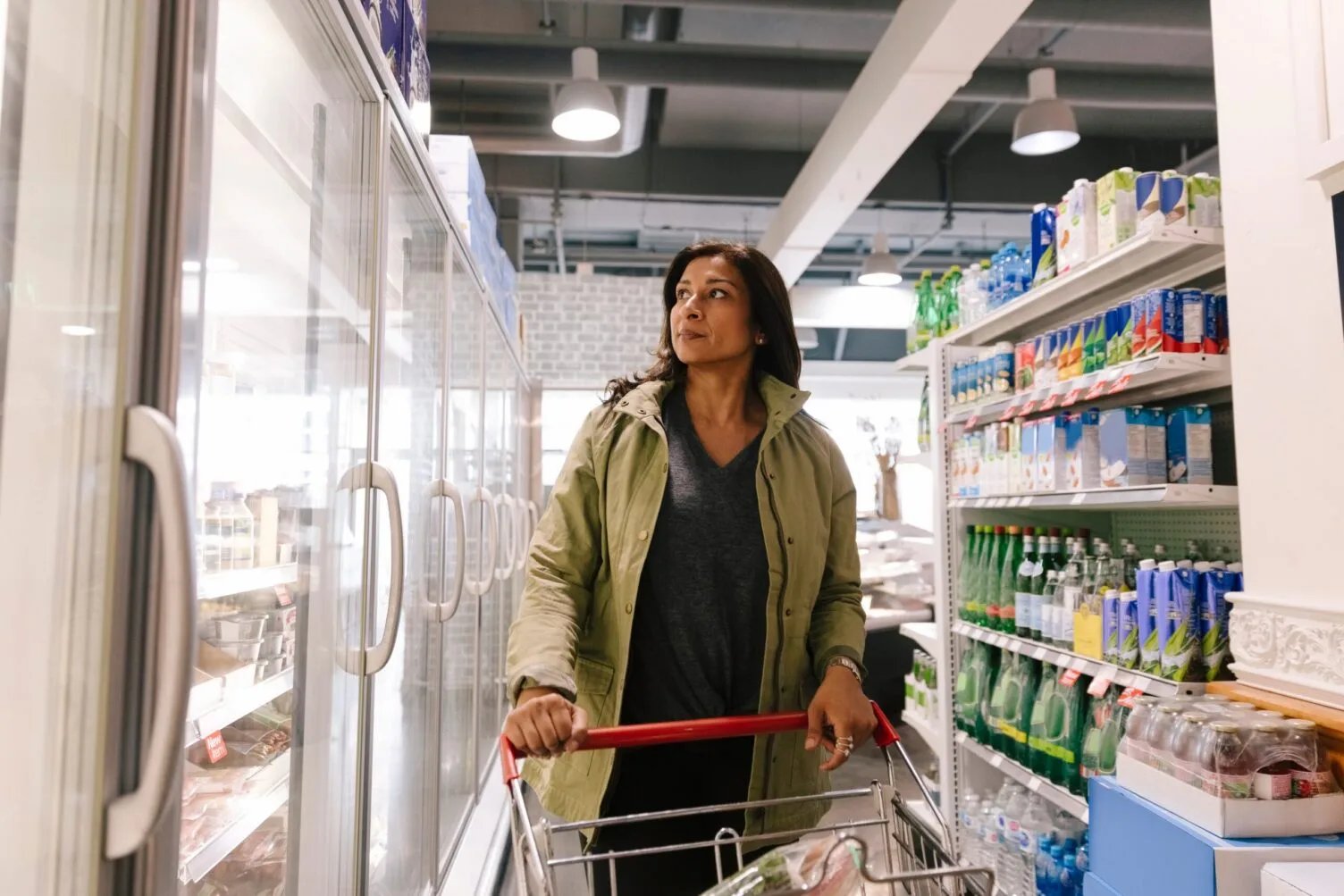 Woman shopping in grocery store dairy aisle with cart, looking at refrigerated products