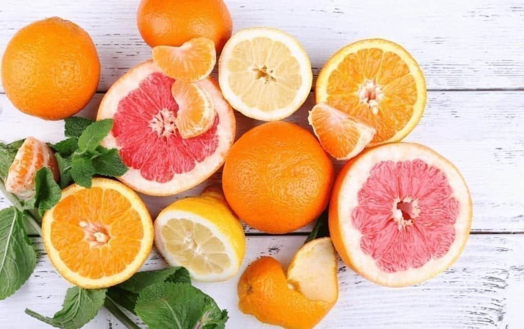 A variety of citrus fruits are displayed on a white wooden surface. Whole and halved oranges, lemons and grapefruits are mixed with peeled segments. There are also green leaves scattered among the fruits, showing the health benefits of citrus in a vivid array. MyFitnessPal blog