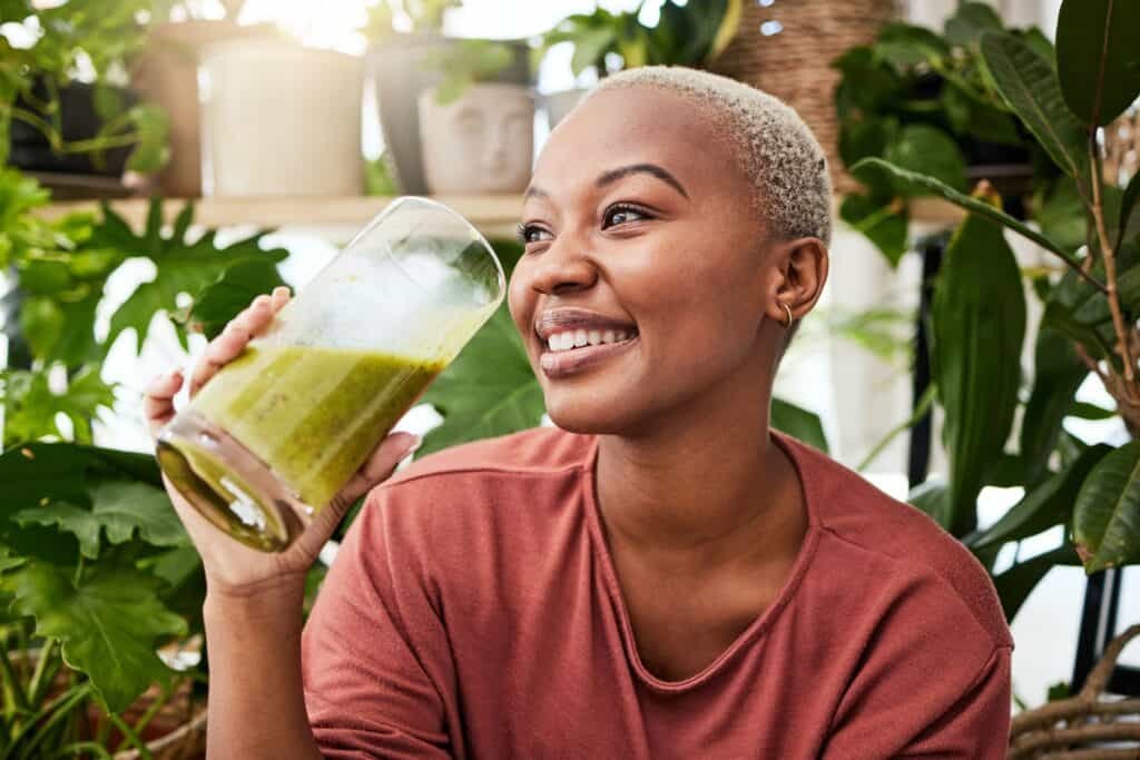 Smiling woman with short blond hair drinking green smoothie surrounded by houseplants