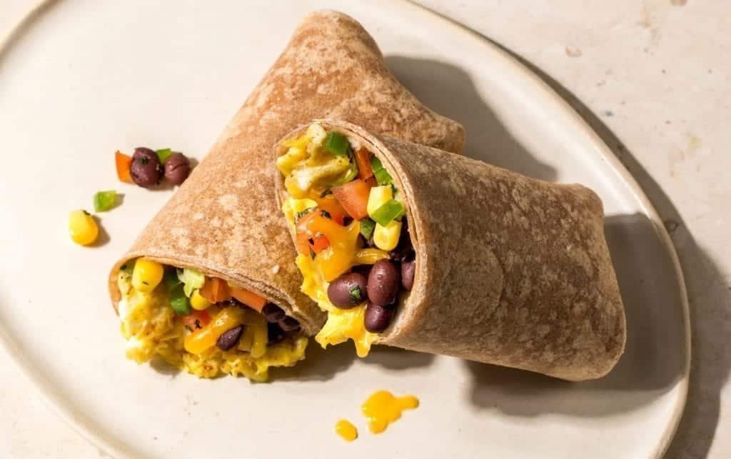 A whole wheat wrap cut in half reveals a filling of scrambled eggs, black beans, chopped tomatoes, corn and jalapeños. These healthy breakfast burritos sit on a round, beige plate on top of a light surface. MyFitnessPal blog