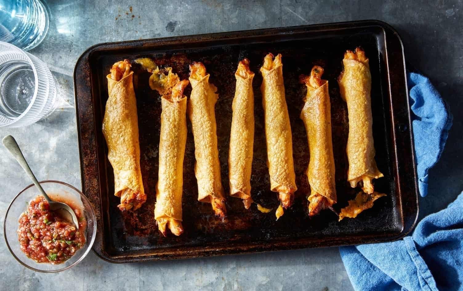 A baking tray with eight crispy chicken taquitos is placed on a metal surface. A bowl of salsa with a spoon beside it and a blue cloth are nearby, along with two glasses of water. The taquitos appear evenly spaced on the tray. MyFitnessPal Blog