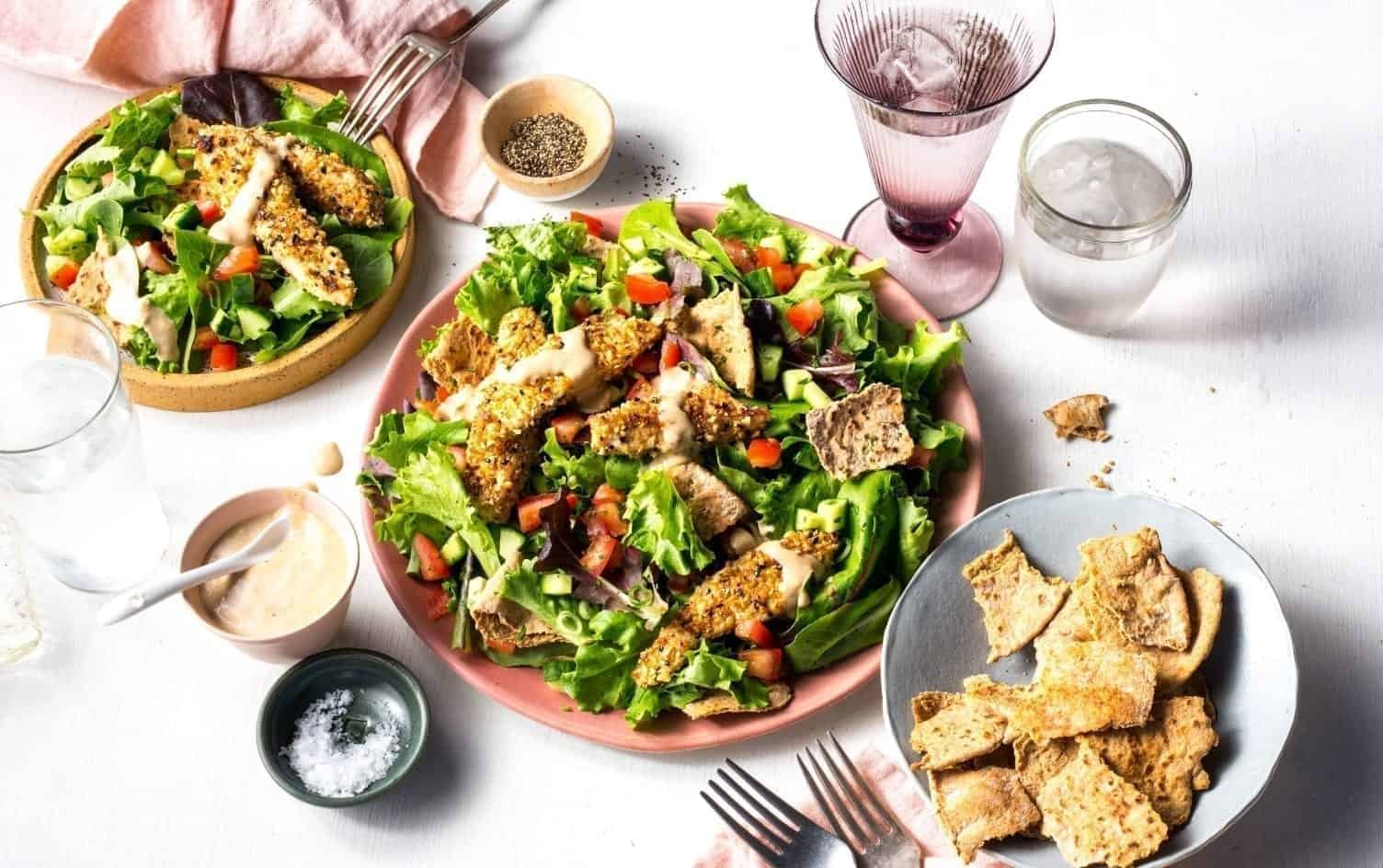 A plate of salad with grilled chicken strips resembling shawarma, lettuce, tomatoes, croutons, and dressing is accompanied by a smaller bowl of salad, a plate of croutons, and three drinks in glasses. A bowl of sauce is on the side with salt and pepper. Cutlery is visible. MyFitnessPal Blog