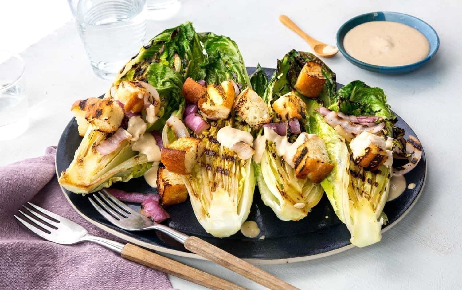 A plate of grilled romaine lettuce topped with croutons, grilled onions, and a creamy cashew dressing. Two forks and a knife are placed near the plate. There is a small bowl of dressing and a glass of water in the background. A cloth napkin lies under the utensils. MyFitnessPal Blog