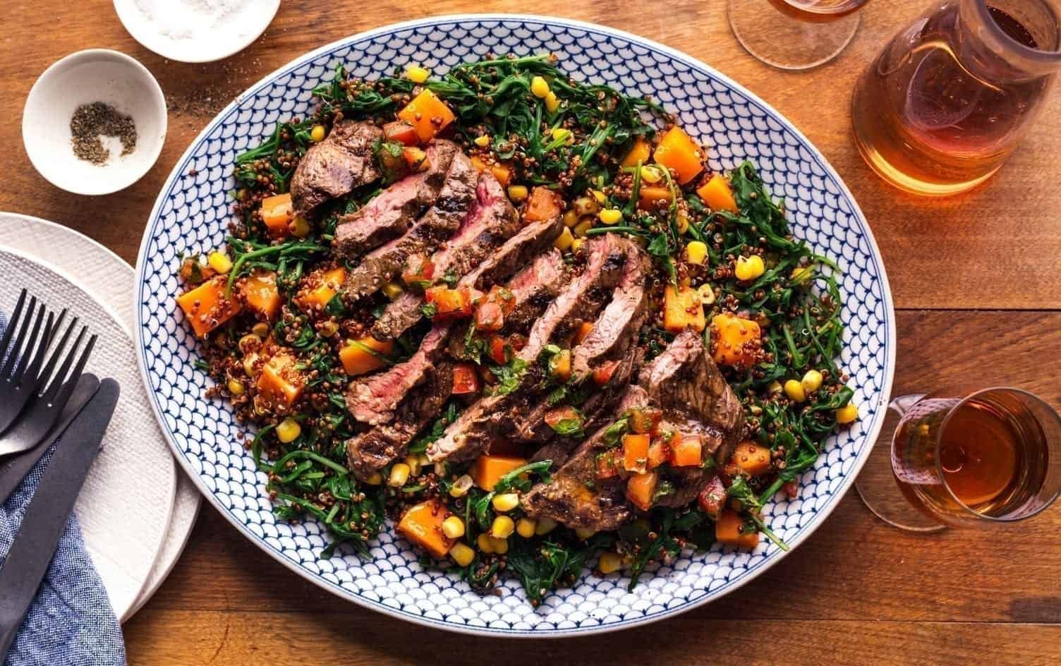 A plate of sliced grilled steak is served on a bed of quinoa, corn, spinach, and diced butternut squash. The dish rests on a wooden table, accompanied by a white bowl of salt and pepper, a glass of amber-colored liquid, and utensils placed neatly on a napkin. MyFitnessPal Blog