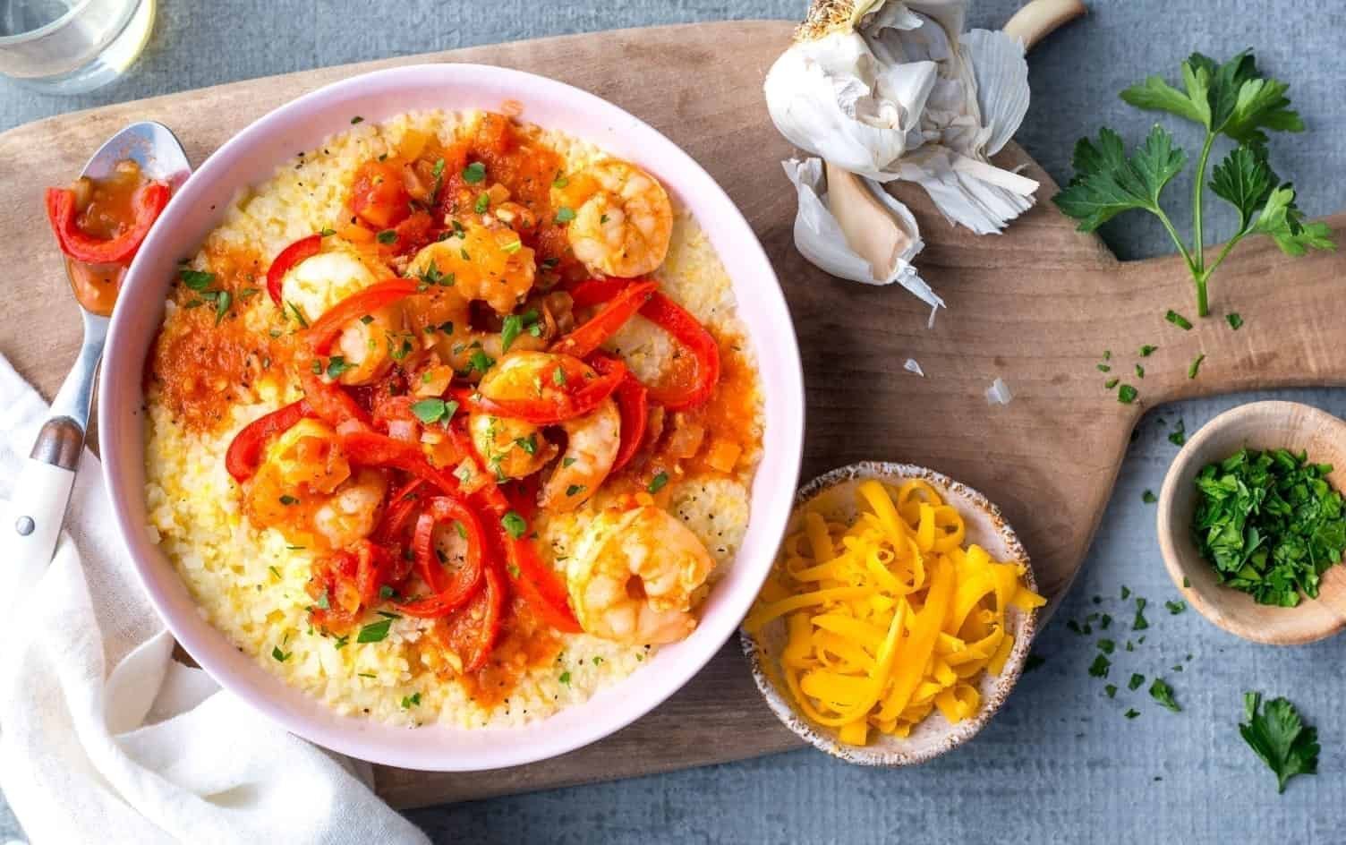 A bowl of shrimp and cauliflower grits is placed on a wooden board. The dish is garnished with red bell pepper slices, herbs, and sauce. Surrounding the bowl are garlic, parsley, a small bowl of shredded cheese, a small bowl of chopped herbs, and a cloth napkin. MyFitnessPal Blog
