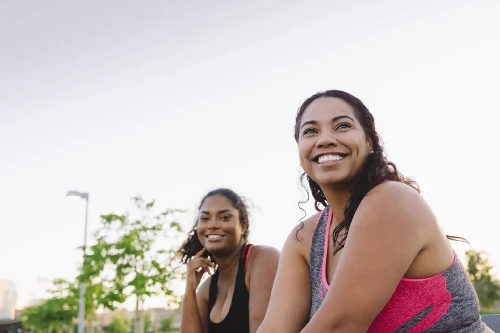 Two smiling women in sportswear outdoors, one in pink sports bra, one in black, enjoying fitness together