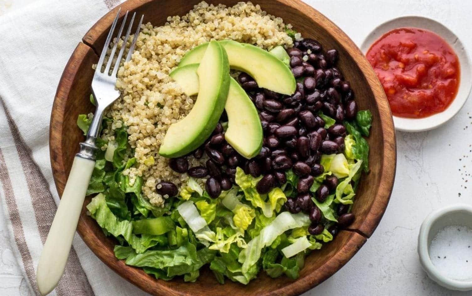 A wooden bowl contains five ingredients: quinoa, black beans, sliced avocado, and chopped lettuce. A fork rests on the side of the bowl. To the right, there is a small dish of red sauce. A beige striped napkin is partially visible underneath the bowl. MyFitnessPal Blog