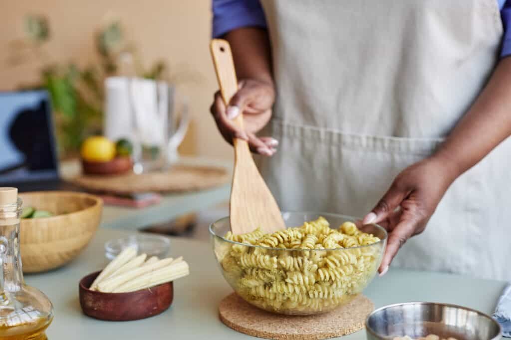 Woman stirring fusilli pasta with pesto in glass bowl while cooking healthy meal at home