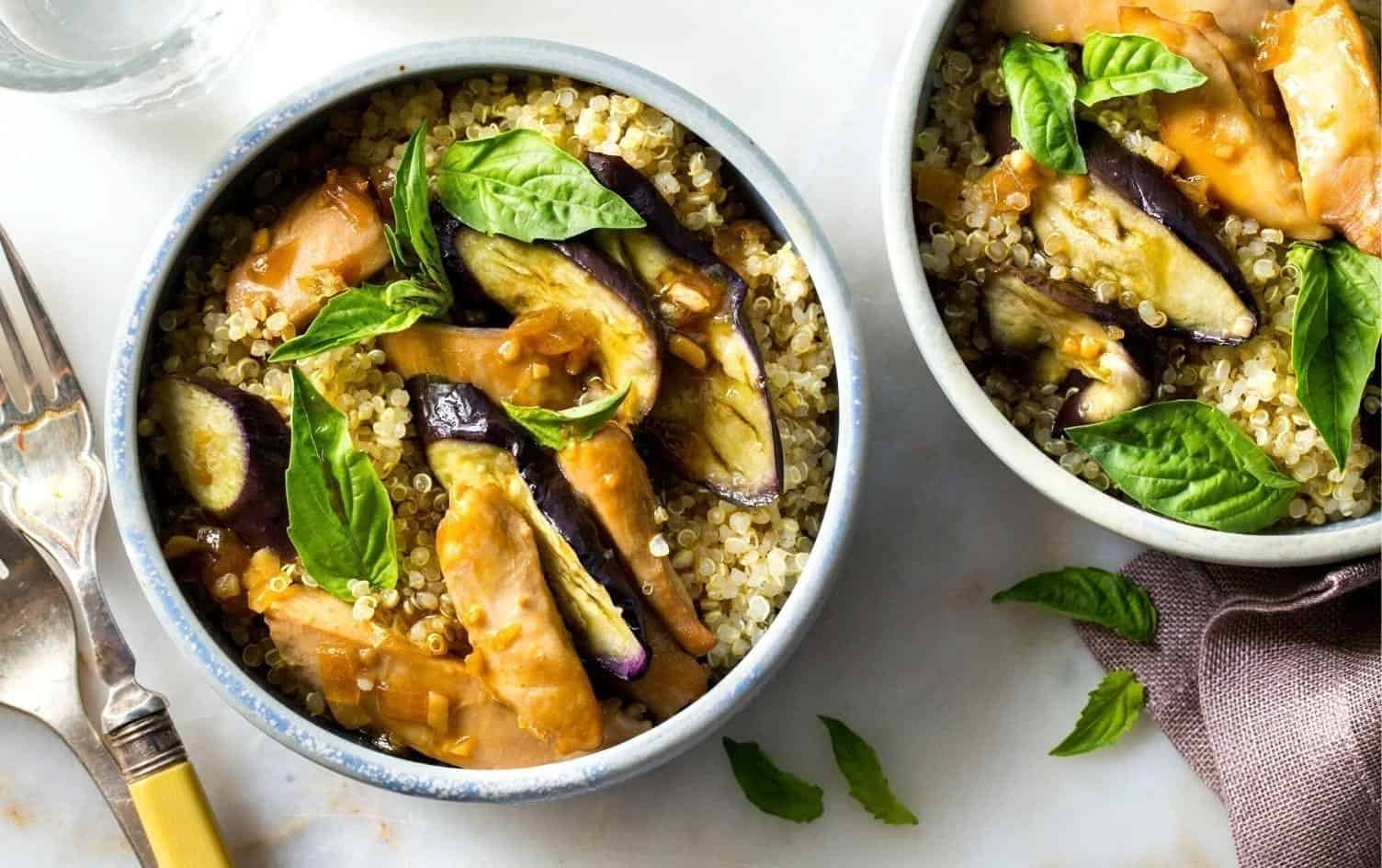 Two bowls of quinoa salad are placed on a white surface. Each bowl contains chunks of instant pot chicken, roasted eggplant slices, fresh basil leaves, and cooked quinoa. A fork and knife are positioned nearby, along with a glass of water. MyFitnessPal Blog