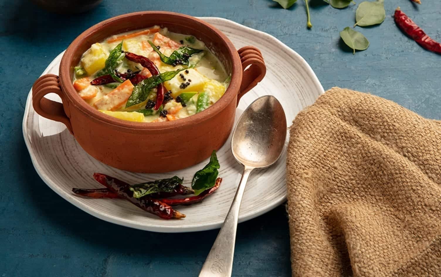 A clay pot filled with colorful vegetable curry, reminiscent of avial, garnished with curry leaves and red chili peppers on a white plate. A silver spoon and a beige cloth napkin are placed next to the dish. The setting is on a blue surface with a few scattered green leaves and red chilies. MyFitnessPal Blog