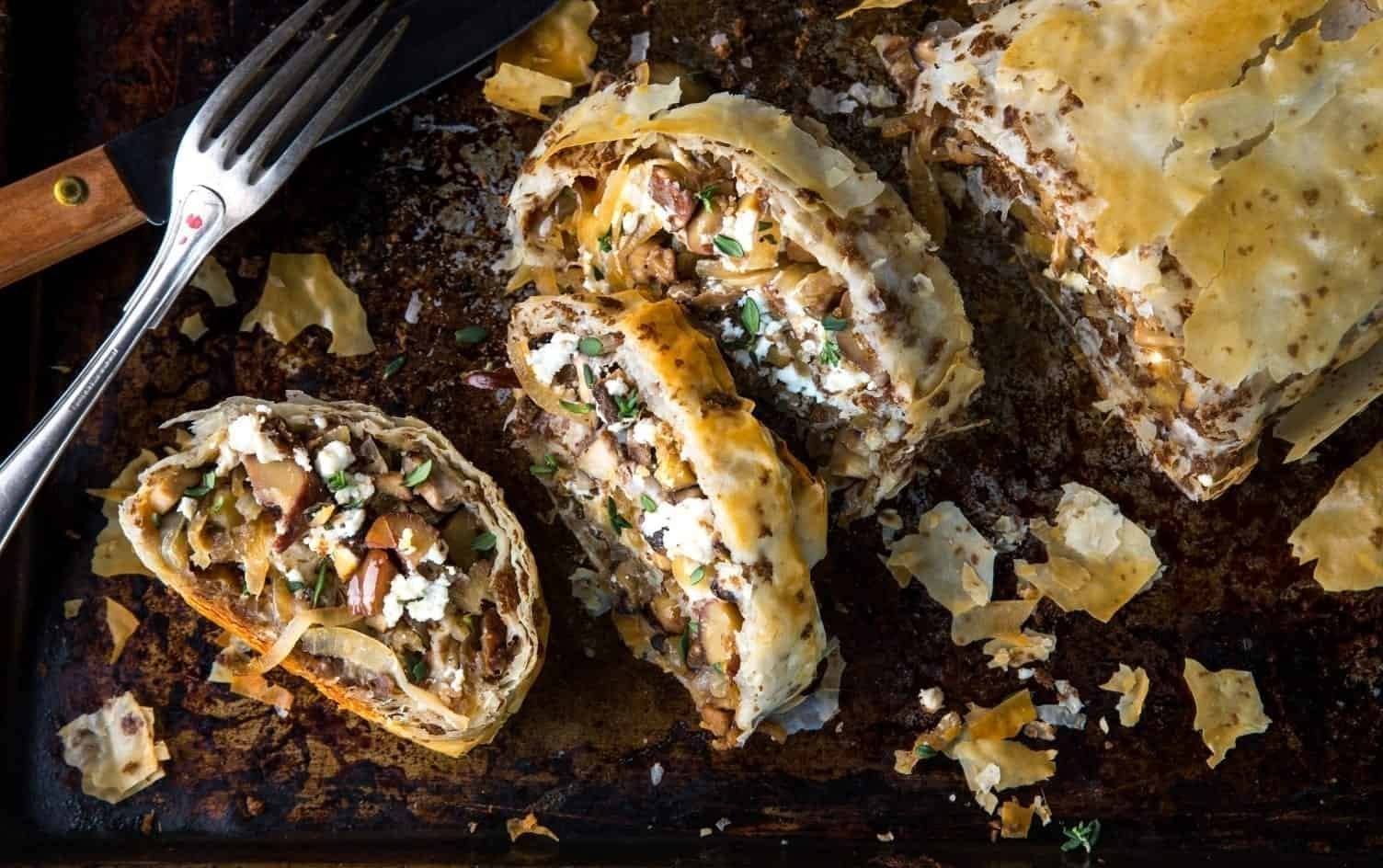 A sliced puff pastry roll filled with a savory mixture of mushrooms, spinach, and cheese is displayed on a dark baking tray. Crumbs and small pieces of the pastry are scattered around, with a fork and knife placed nearby. MyFitnessPal Blog