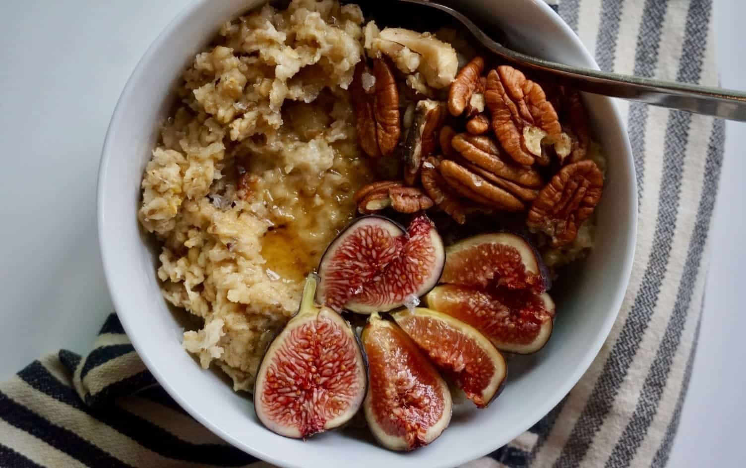 A bowl of instant pot oats topped with halved fresh figs and pecans. A silver spoon rests inside the bowl, which is placed on a white-and-black striped cloth against a light gray background. MyFitnessPal Blog