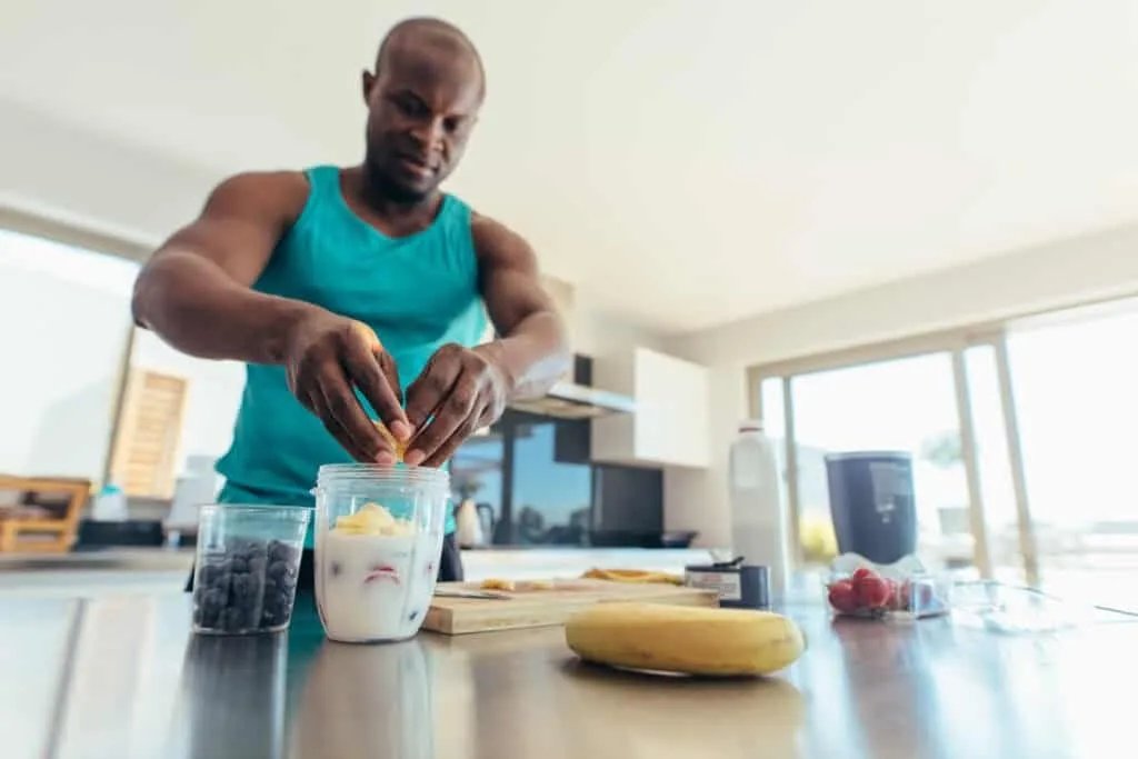 Athletic man in turquoise tank top making protein smoothie with banana and berries in modern kitchen
