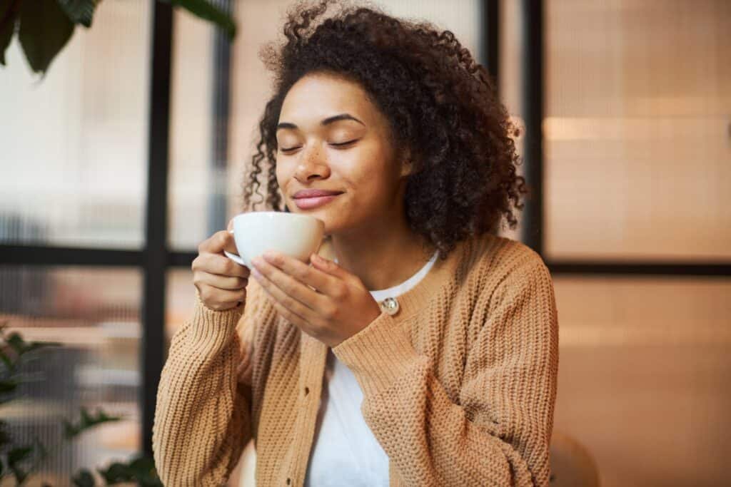 Woman with curly hair enjoying coffee with closed eyes in bright cafe