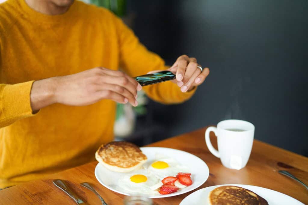 Person in yellow sweater photographs breakfast with pancakes, fried eggs and strawberries using smartphone
