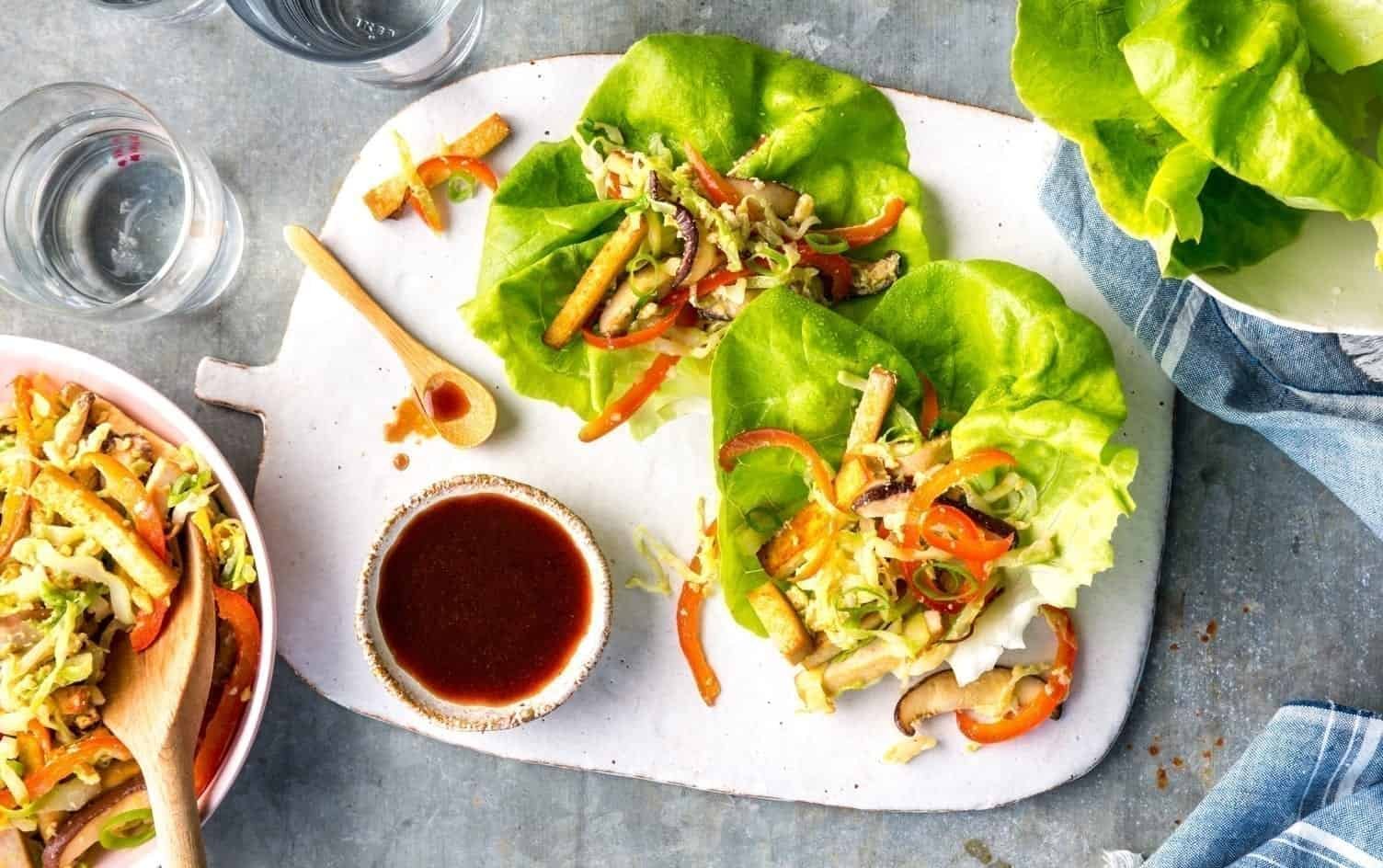 A white cutting board on a grey surface holds two lettuce wraps filled with vegetables and tofu. A small bowl of sauce with a wooden spoon is nearby. In the background, there's a bowl of more vegetable filling and tofu, a plate with lettuce leaves, and a blue cloth napkin. MyFitnessPal Blog