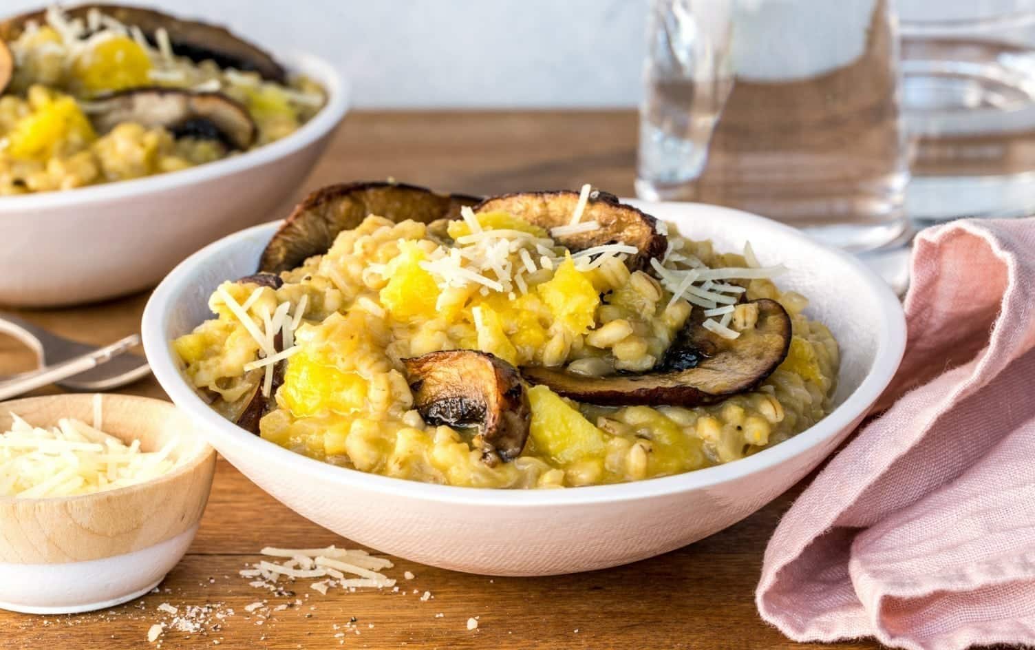 A bowl of risotto topped with grilled mushrooms, chunks of pumpkin, and grated cheese sits on a wooden table next to a smaller bowl of grated cheese, a pink napkin, and a glass of water. Another enticing bowl of risotto is in the background. MyFitnessPal Blog